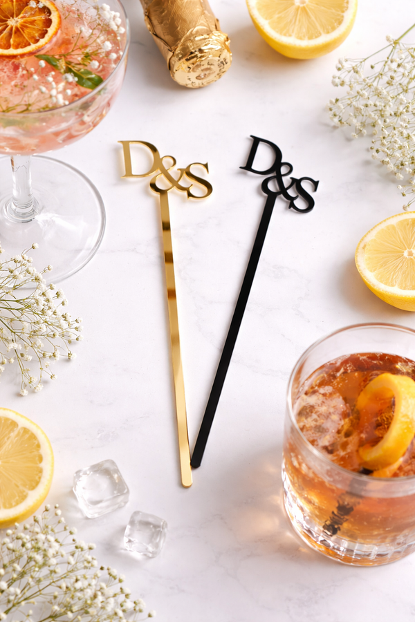 Two cocktail stirrers with 'D&S' initials next to glasses of cocktails with lemon slices on a white surface.