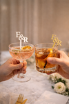 Two hands holding cocktails with 'E&R' stirrers against a blurred background